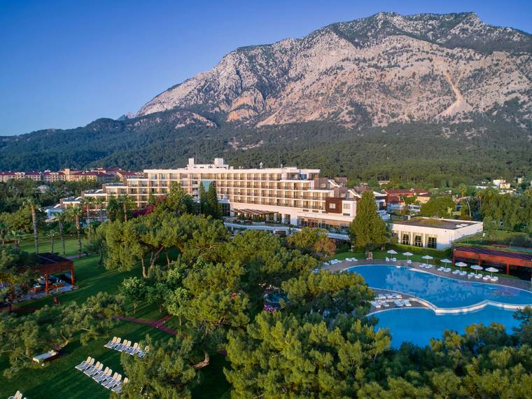 Aerial view of the Rixos Beldibi resort complex nestled between the Mediterranean Sea and the steep, forested mountains of Antalya, Turkey. The multi-story hotel building with numerous balconies is surrounded by lush green lawns and tall trees. In the foreground, there is a large, irregularly shaped outdoor swimming pool with sun loungers and umbrellas.