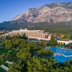 Aerial view of the Rixos Beldibi resort complex nestled between the Mediterranean Sea and the steep, forested mountains of Antalya, Turkey. The multi-story hotel building with numerous balconies is surrounded by lush green lawns and tall trees. In the foreground, there is a large, irregularly shaped outdoor swimming pool with sun loungers and umbrellas.